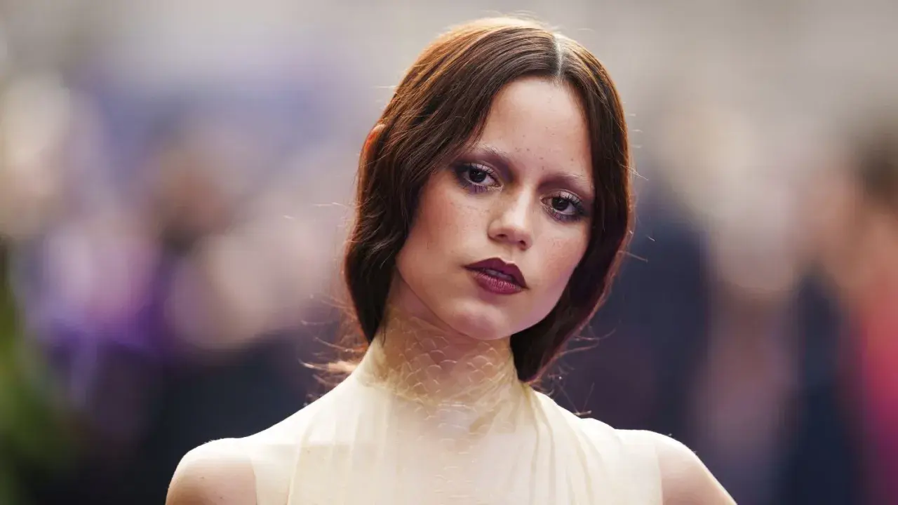 A young woman with auburn hair and a dramatic, dark lipstick looks directly at the camera. Her eye makeup is smudged, giving her a tired girl makeup look, as if she's been out all night.