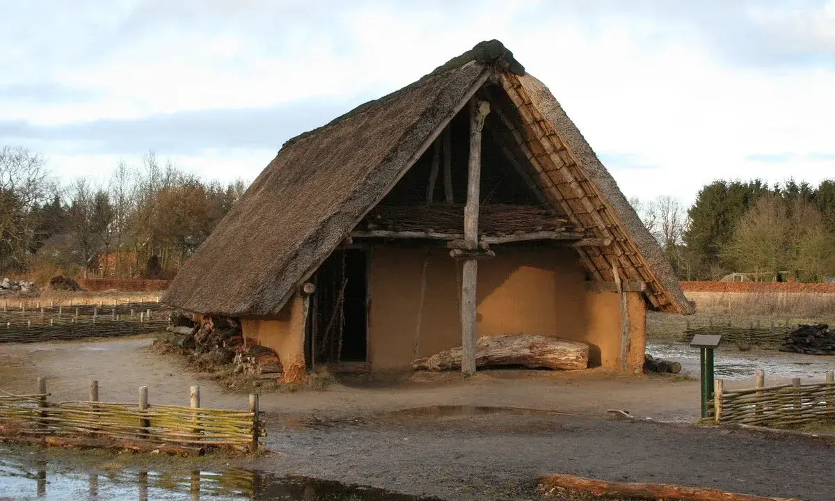 Ein rustikales Bauernhaus aus dem Mittelalter mit Strohdach und Lehmwänden, umgeben von Holzzäunen und Bäumen.