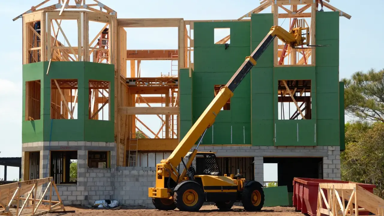 Chantier d'une maison individuelle en cours de construction. Une grue télescopique jaune est positionnée près de la structure en bois et des panneaux verts.