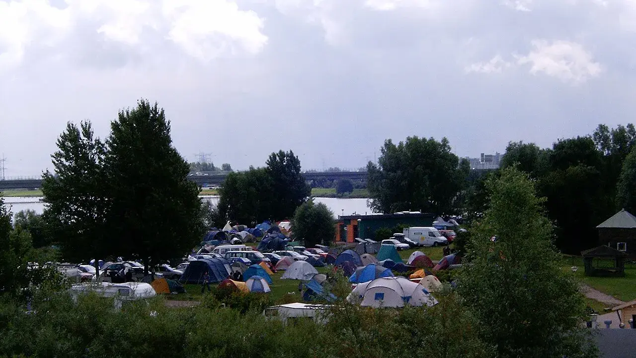Rozległy kamping z mnóstwem namiotów i samochodów, nad wodą, pod pochmurnym niebem.