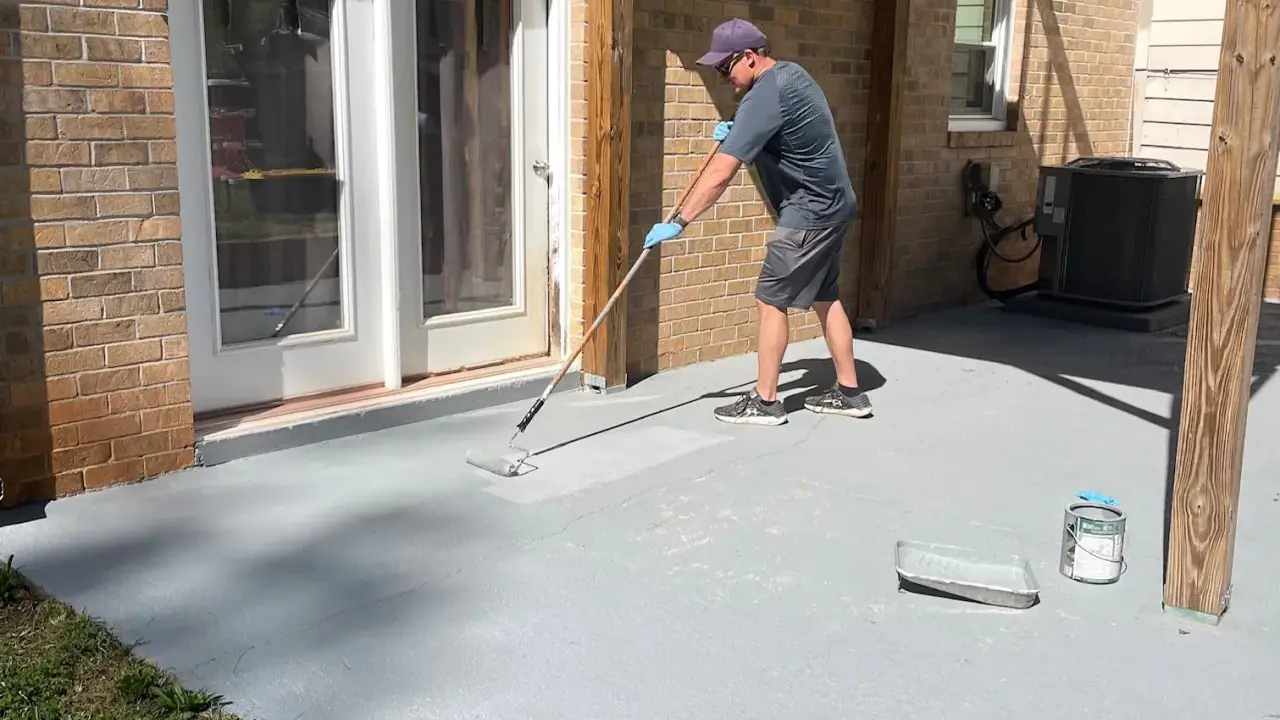 Un homme applique une peinture sur mur béton extérieur avec un rouleau. Le sol gris est en cours de rénovation.