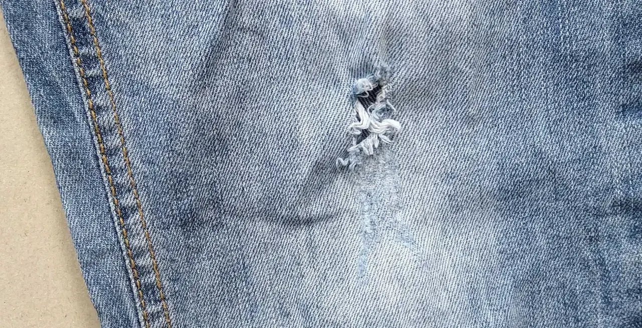 Jak zaszyć dziurę w spodniach dresowych? Widok z bliska na przetarty jeans z widocznym uszkodzeniem, które można naprawić.