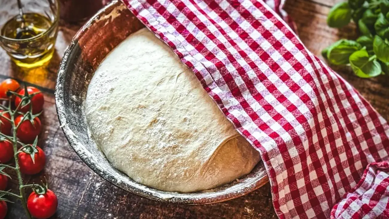 Frischer Pizzateig ruht in einer Schüssel, bedeckt mit einem rot-weiß karierten Tuch. Die Frage: wie viel Hefe in Pizzateig?