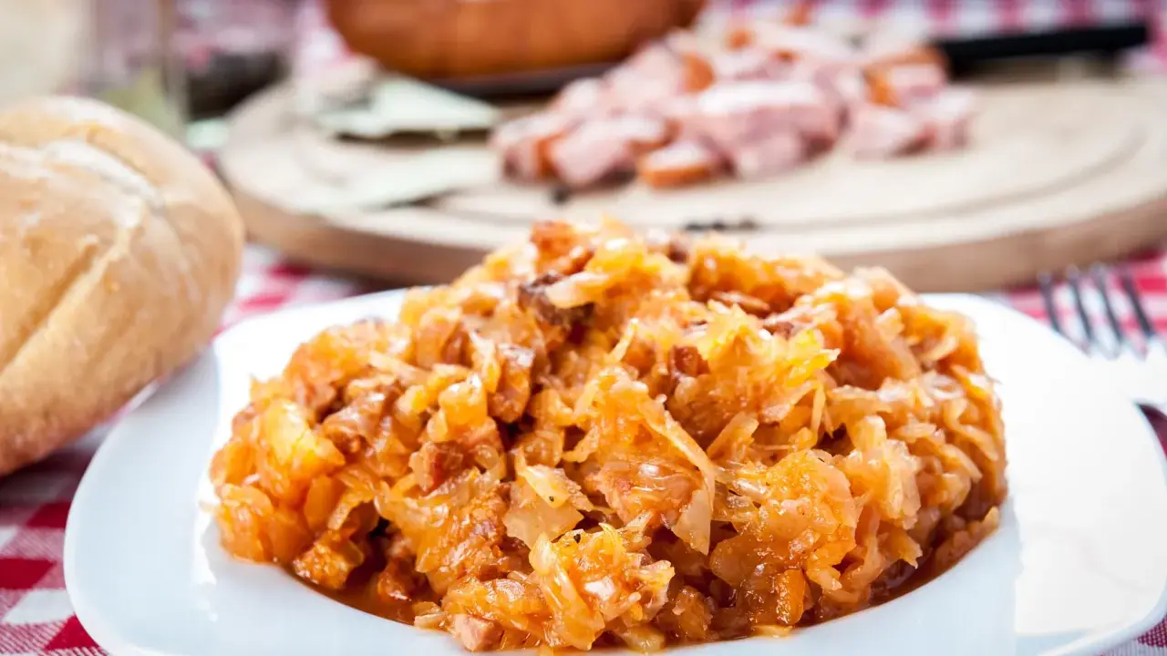Pyszny bigos Ani, gotowany z pasją, na talerzu. Obok bułka i kawałki kiełbasy.