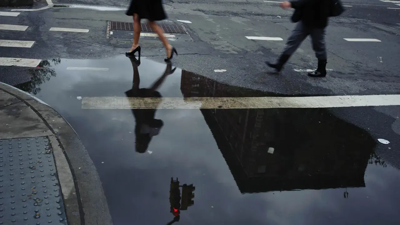 Jay Maisel photography captures a rainy street scene with blurred figures crossing, their reflections distorted in puddles, mirroring the city's mood.