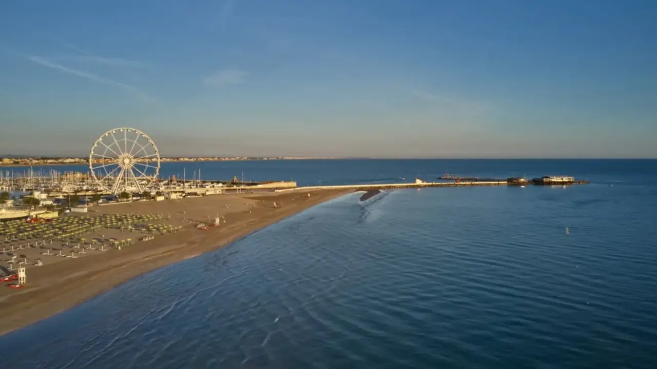 Vista aerea di una spiaggia di Rimini con ruota panoramica, porto turistico e mare calmo sotto un cielo azzurro.
