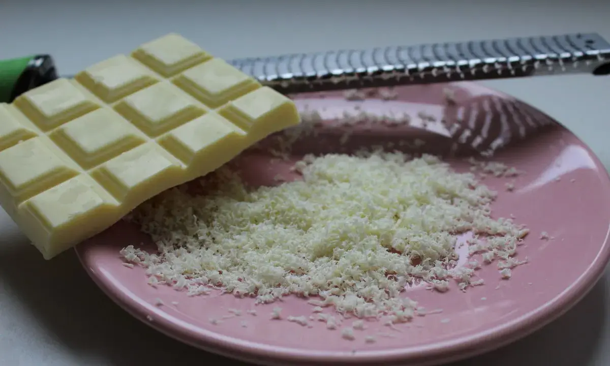 Une râpe à fromage argentée est posée sur une assiette rose, recouverte de copeaux de chocolat blanc fraîchement râpé.