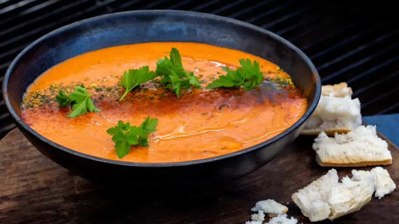 En skål med krämig, orange soppa, troligen en smakrik mojo rojo, garnerad med persilja och serverad med brödbitar.
