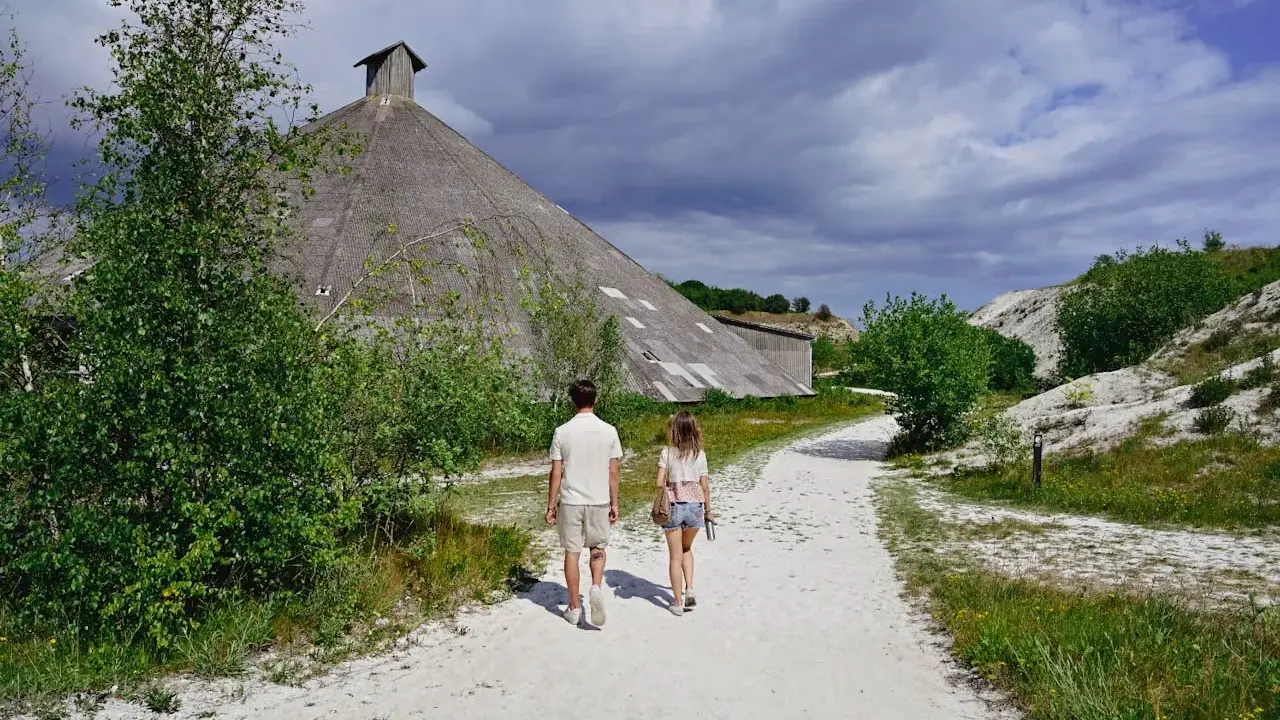 Två personer går på en grusväg mot en stor, konformad byggnad. Omgivningen påminner om vita klippor i Danmark, med grönska och en dramatisk himmel.