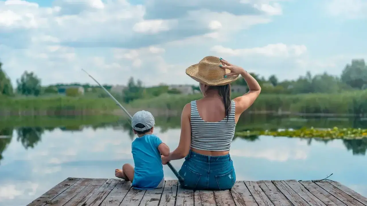 Matka i syn siedzą na drewnianym pomoście, łowiąc ryby. Spokojne łowiska wędkarskie, odbijające się w wodzie chmury.