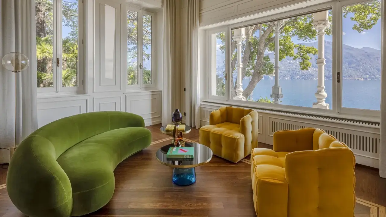 Salotto con divano verde curvo e poltrone gialle, tavolini di design e vista lago. Un'elegante casa Art Déco.