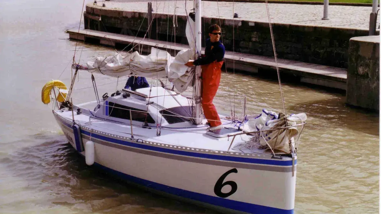 Un homme en salopette rouge prépare un voilier coco, le numéro 6 bien visible sur la proue, prêt à naviguer sur un canal.