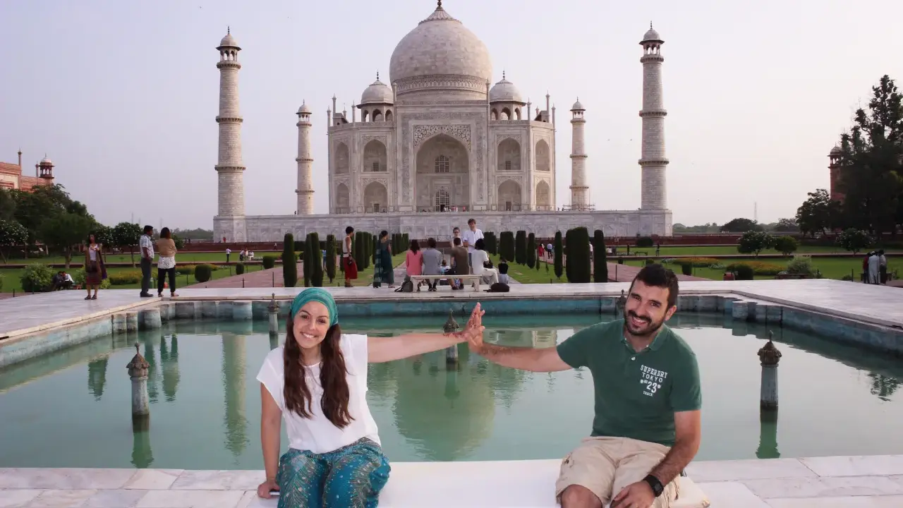 Pareja sonriente en la India, frente al Taj Mahal, con niños y visitantes al fondo.