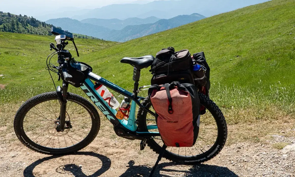 Bereit für e bike touren: Ein Trek E-Bike mit Gepäcktaschen steht auf einem Schotterweg, umgeben von grünen Hügeln und Bergketten.