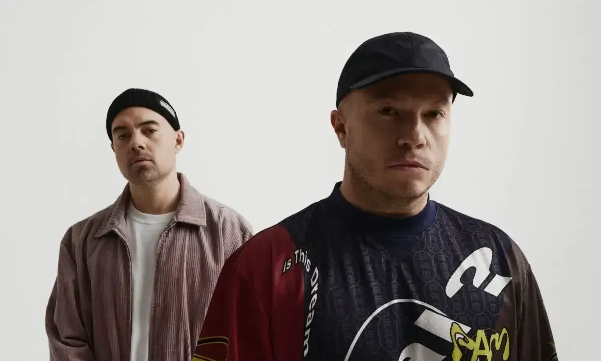 Dwóch mężczyzn, jeden w czapce beanie, drugi w czapce z daszkiem. Na koszulce jednego z nich widoczny jest **guzior fala tekst**.