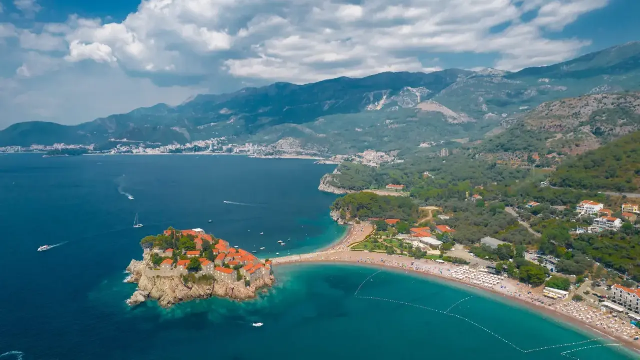 Malownicza wyspa Sveti Stefan, połączona z lądem piaszczystą mierzeją, otoczona turkusowym morzem i górami.