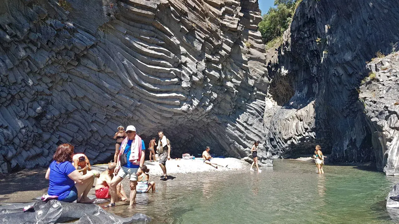 Persone si rilassano e si rinfrescano nelle acque cristalline delle cascate dell'Alcantara, circondate da imponenti pareti rocciose basaltiche.