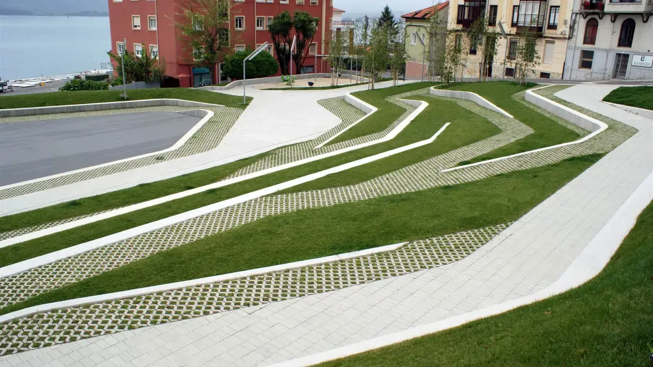 Diseño moderno con césped y caminos de hormigón que forman patrones ondulados. El suelo drenante permite el paso del agua, integrándose con el entorno urbano y el mar al fondo.