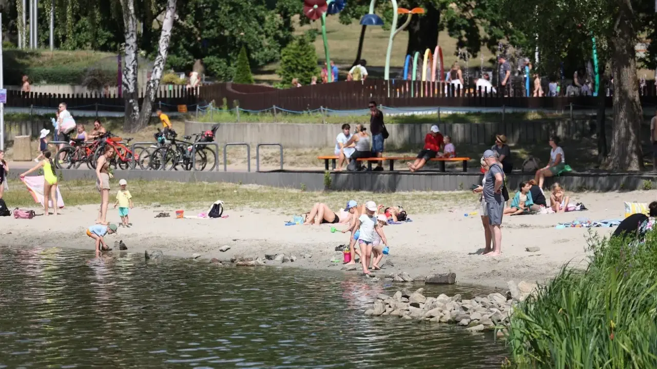 Rodzina odpoczywa nad wodą na piaszczystej plaży Jeziora Paprocany. Dzieci bawią się w piasku i przy brzegu, dorośli relaksują się na kocach i ławkach.