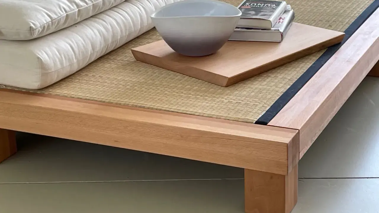 Eine gestapelte, weiße Matratze eines japanischen Bettes ruht auf einer Tatami-Matte. Daneben liegen ein Buch und eine Schale auf einem Holzbrett.