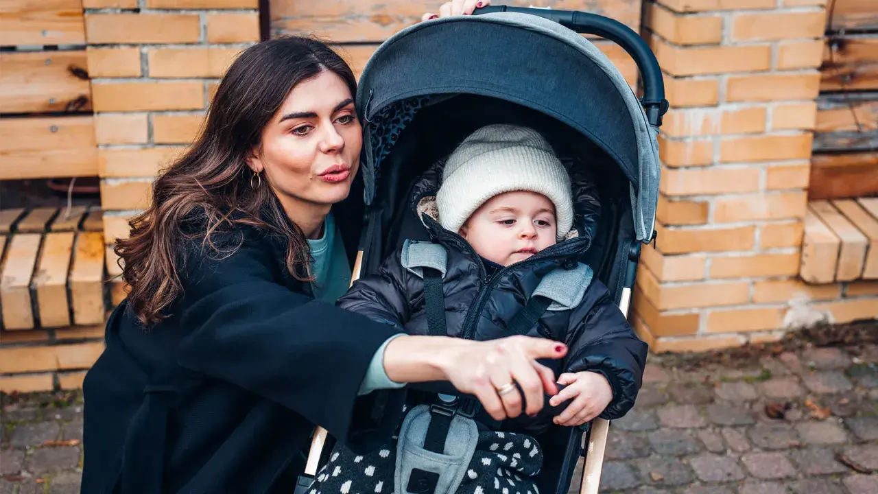 Mama pokazuje coś dziecku w najmniejszej spacerówce. Maluch w czapce i kurtce wygląda ciekawie.