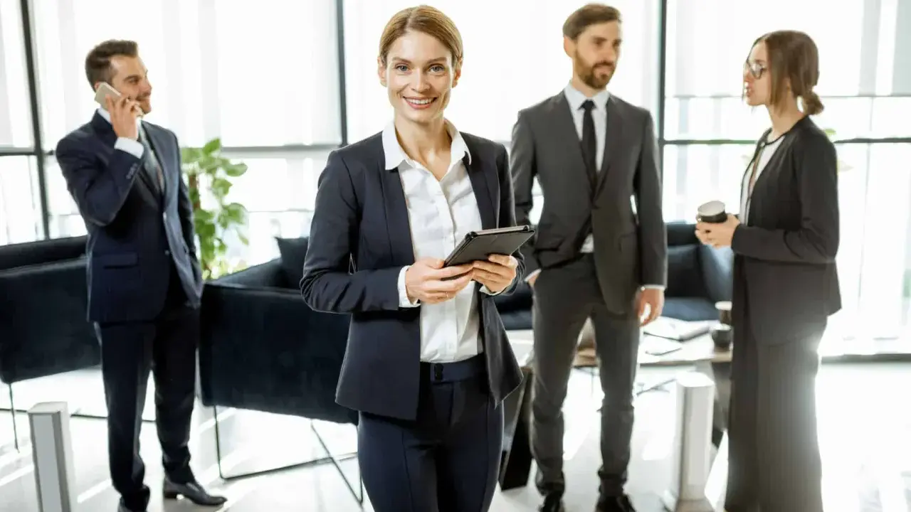 Une femme souriante, possiblement le chief of staff, tient une tablette. Autour d'elle, des collègues en costume discutent dans un bureau moderne.
