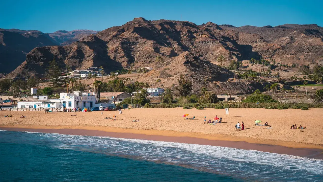 Słoneczne gran canaria plaże z piaszczystym brzegiem, turkusową wodą i górami w tle. Ludzie odpoczywają pod parasolami.