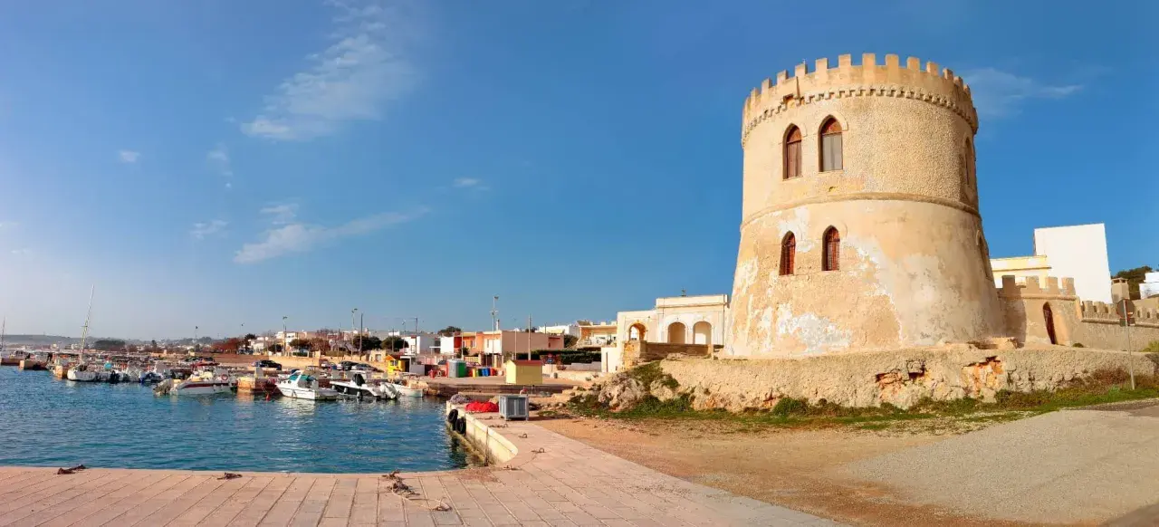 Torre Vado, Lecce: un'antica torre di avvistamento domina un porto con barche e case colorate sotto un cielo azzurro.