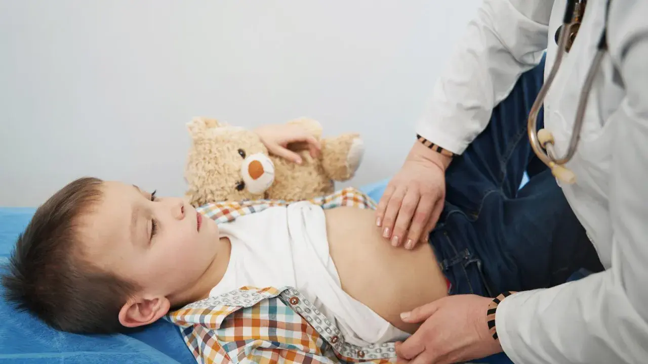 Lekarz bada brzuch dziecka, które skarży się na ból brzucha w okolicy pępka. Obok leży pluszowy miś.