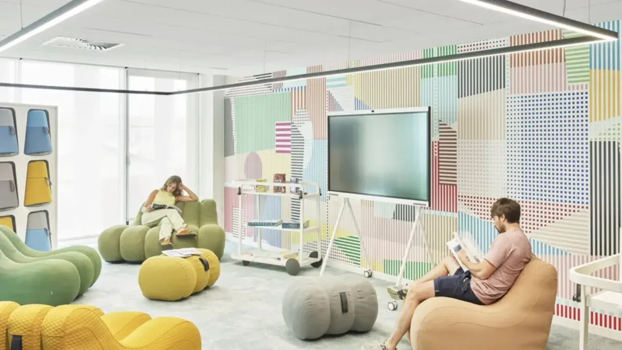 Un espace de repos coloré avec des poufs confortables et un mur à motifs géométriques. Deux personnes lisent tranquillement.
