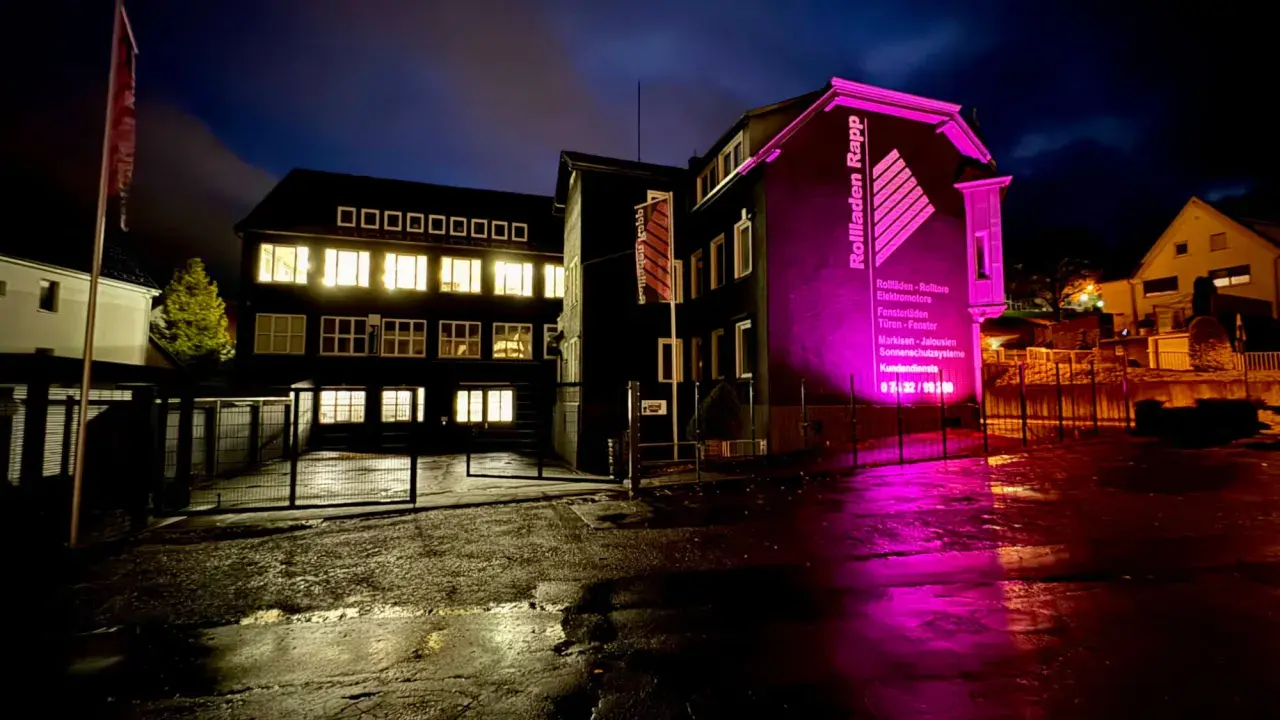 Gebäude mit leuchtenden Fenstern und pinker Beleuchtung. "Rolladen Rapp" wirbt für Nothandkurbel Rolladen, Pflicht für Sicherheit.