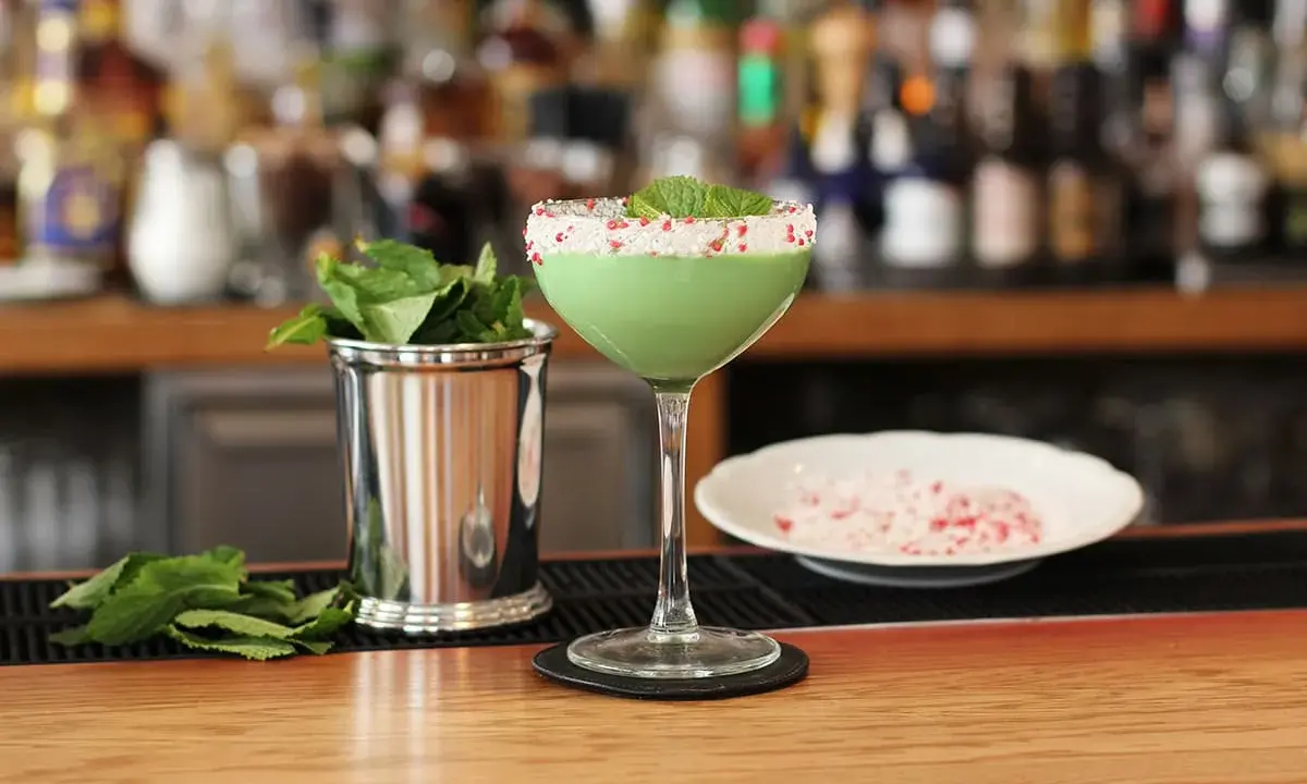 Grön, krämig drink med mynta och krossat strössel på glaskanten. Bredvid står en metallmugg med myntablad och en skål med krossat strössel. Kanske en Amarula drink?