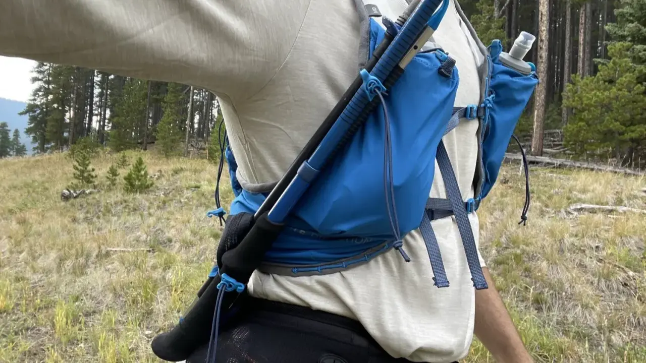 Jak przypiąć kijki do plecaka? Widok z boku na turystę w lesie, z kijami trekkingowymi przymocowanymi do niebieskiego plecaka.