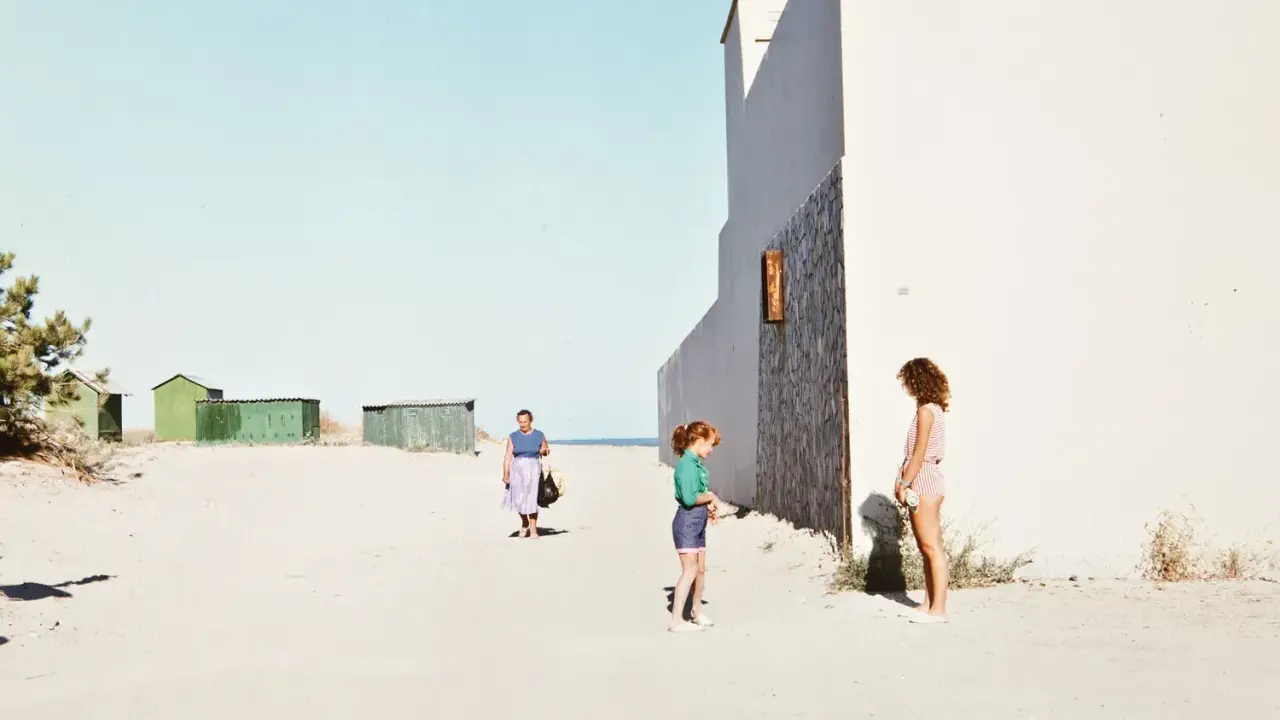 Luigi Ghirri foto: una donna cammina sulla spiaggia, due bambine giocano vicino a un edificio bianco.
