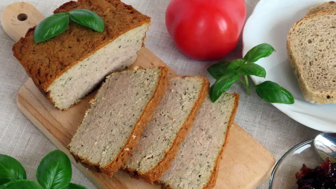 Pokrojony pasztet mieszany drobiowo-wieprzowy na desce, obok pomidor, chleb i konfitura.