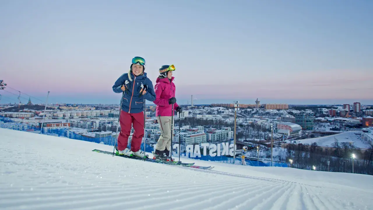 Två skidåkare njuter av utsikten över Hammarbybacken och staden i skymningen.