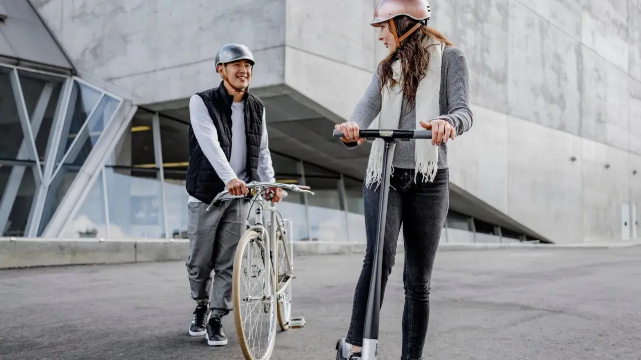 Un homme à vélo et une femme en trottinette, tous deux portant un casque obligatoire, discutent devant un bâtiment moderne.