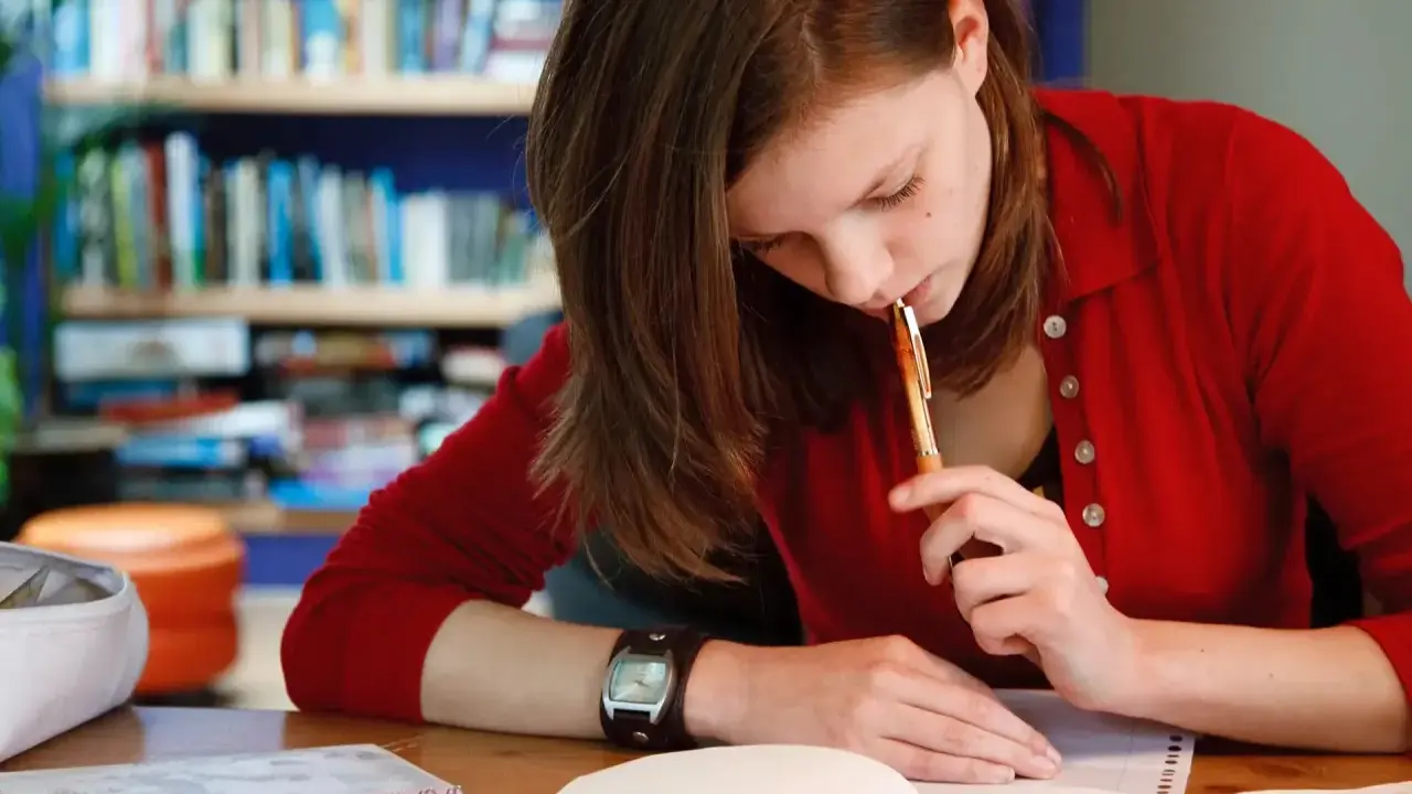 Dziewczyna w czerwonym swetrze myśli nad tekstem rozprawki maturalnej, trzymając długopis przy ustach.