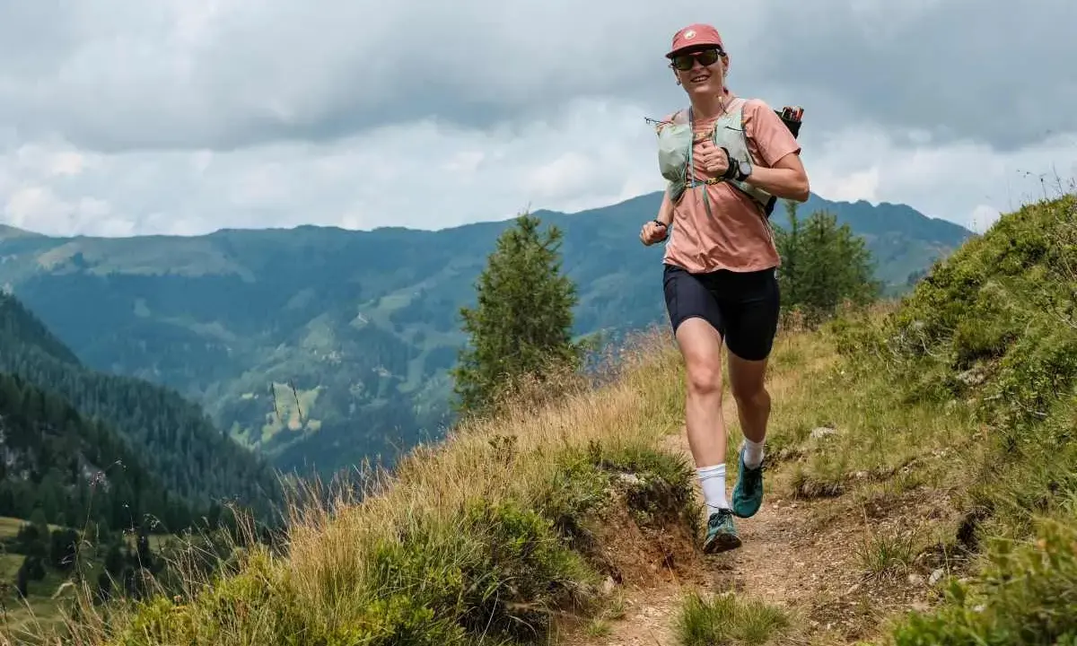 Lächelnde Frau beim Trailrunning Training auf einem Bergpfad, mit Blick auf grüne Täler und bewölkten Himmel.