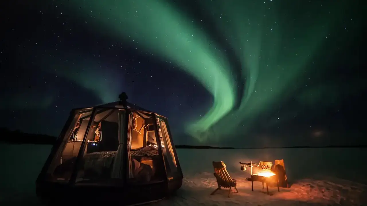 En glaskupol för camping i Norrland lyser upp natten under ett dansande norrsken. En eld sprakar vid sidan om.