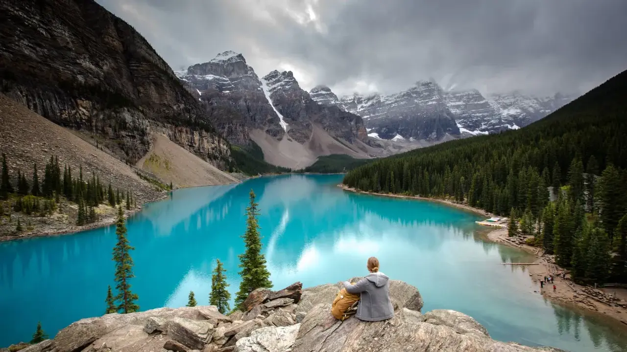 Person sitzt auf Felsen und blickt auf den türkisfarbenen Moränen-See im Banff National Park, Kanada. Majestätische Berge und Nadelwälder umgeben das Wasser.