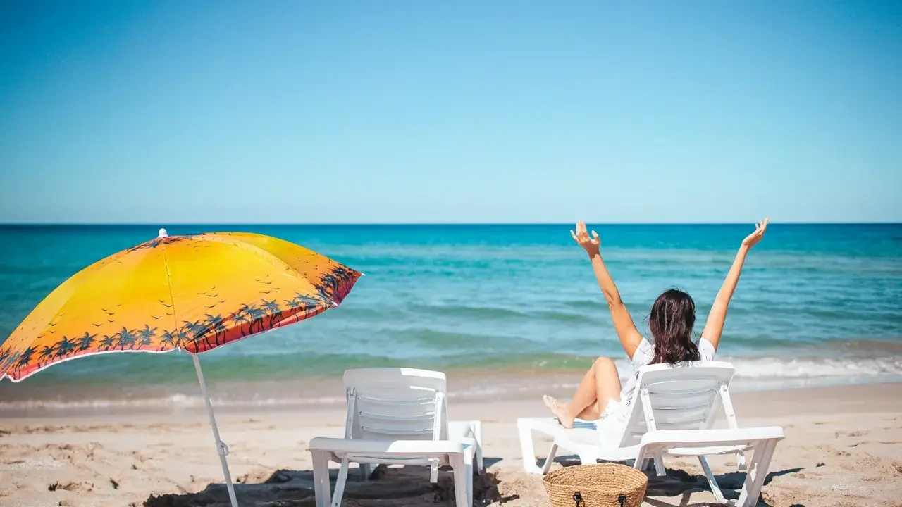 Kobieta cieszy się urlopem wypoczynkowym na plaży, z rękami w górze. Obok leżaki i parasol.