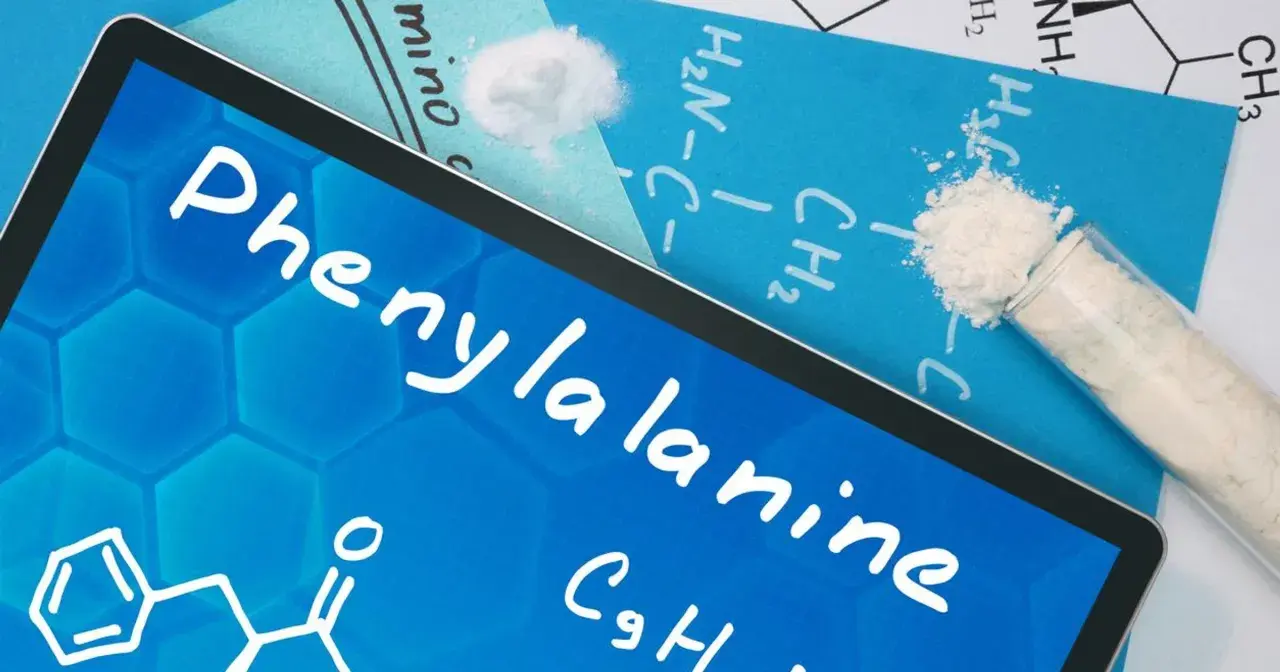 Na ekranie tabletu nazwa "Phenylalanine" i wzór chemiczny. Obok proszek fenyloalaniny w probówce i na kartce.