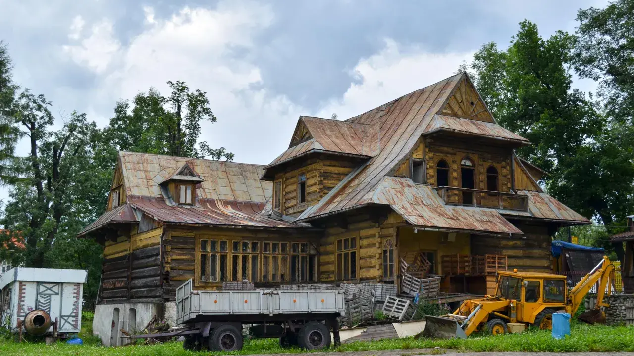 Drewniana chata w Bukowinie Tatrzańskiej, przypominająca starą turnię, z żółtą koparką i przyczepą na podwórzu.