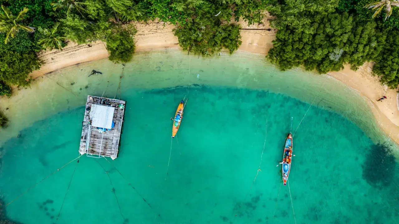 Drohne über Thailand: Ein Blick von oben auf eine schwimmende Plattform und zwei Boote im türkisfarbenen Meer, umgeben von üppiger Vegetation und Sandstrand.