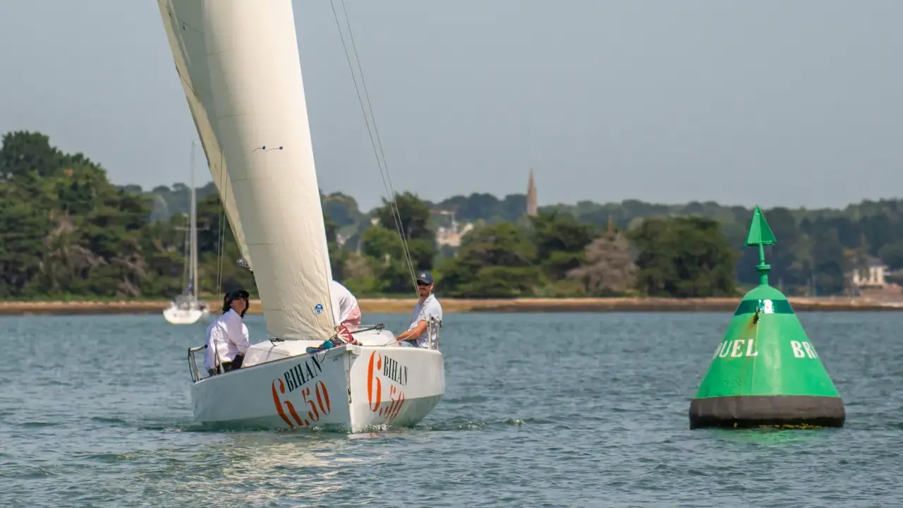 Voile blanche sur l'eau, le voilier "Bihan 6.50" navigue avec deux personnes à bord. Une bouée verte marque le passage.