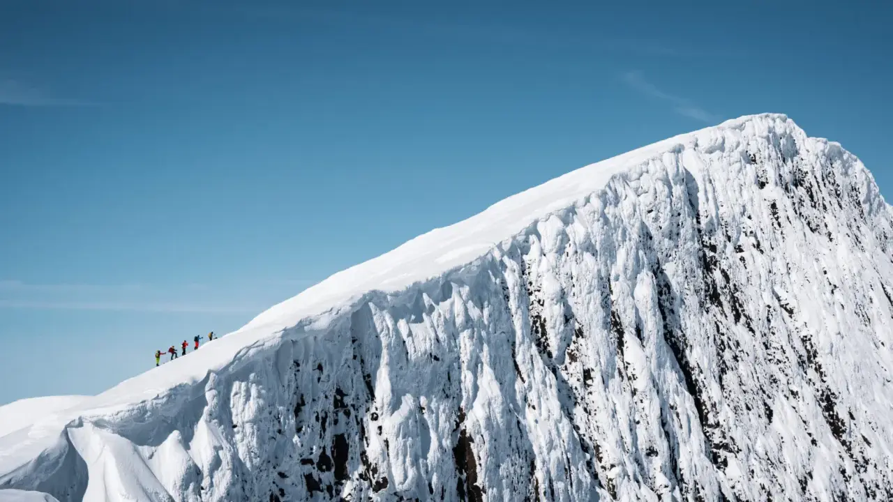 En grupp skidåkare på en helags topptur, med en dramatisk snöklädd bergssida och klarblå himmel.