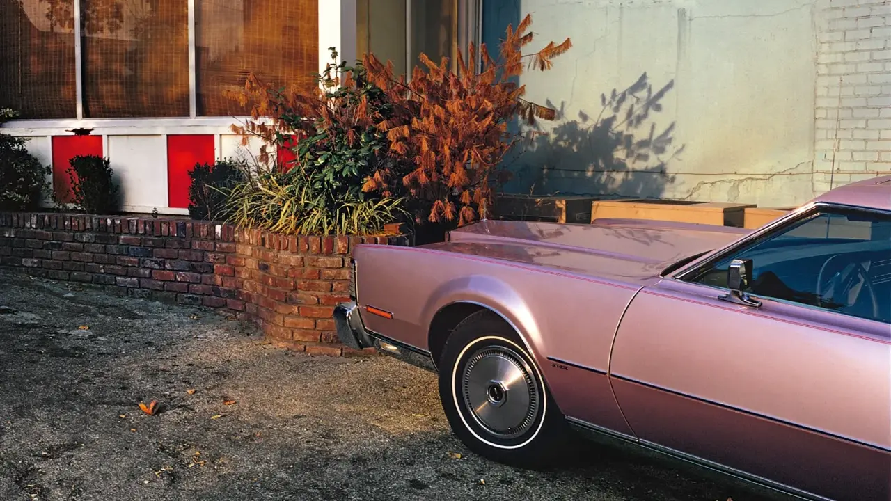 Un'auto rosa pastello, un classico stile William Eggleston, parcheggiata vicino a un muro di mattoni e un edificio con pannelli rossi e bianchi.