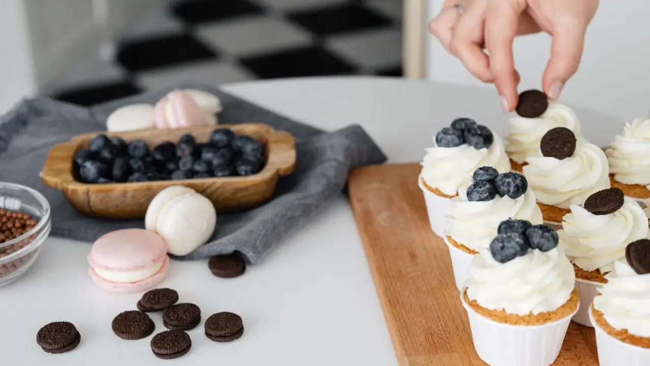 Des cupcakes décorés avec une crème chiboust aérienne, des myrtilles et des biscuits. Des macarons et des myrtilles sont disposés à côté.