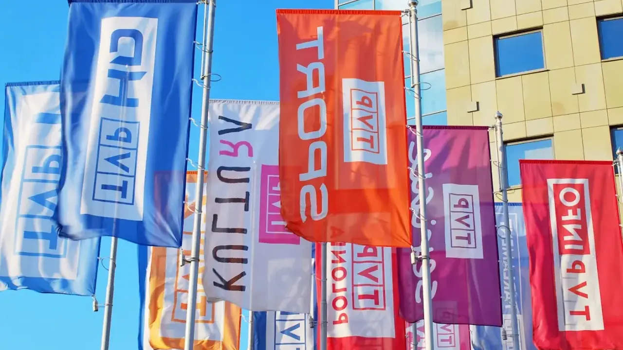 Kolorowe flagi z napisem "TVP w likwidacji" powiewają przed nowoczesnym budynkiem.